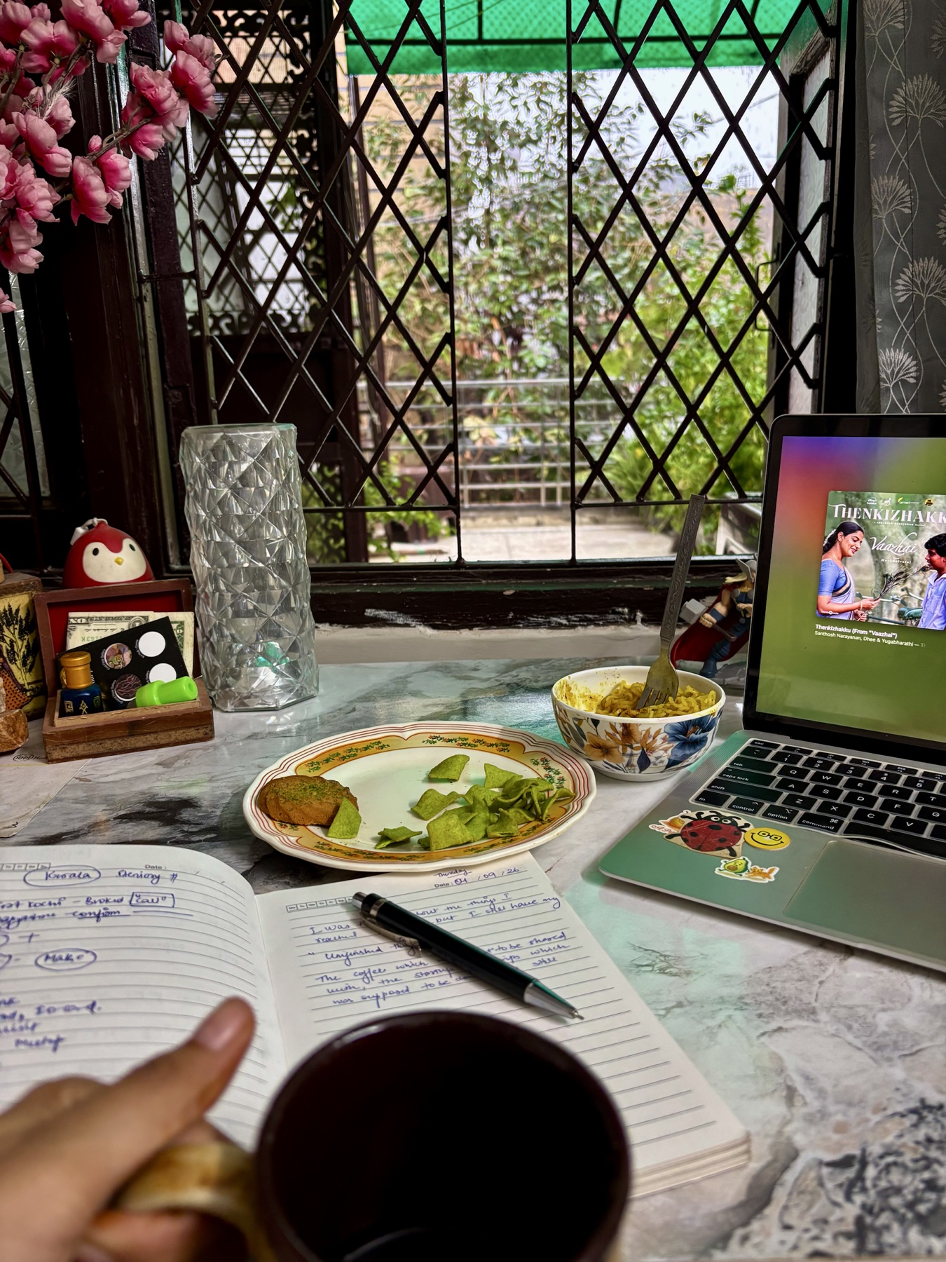A lived-in desk beside a window with half-finished Maggi, an open packet of munchies, coffee, handwritten notes, and a movie playing on a laptop